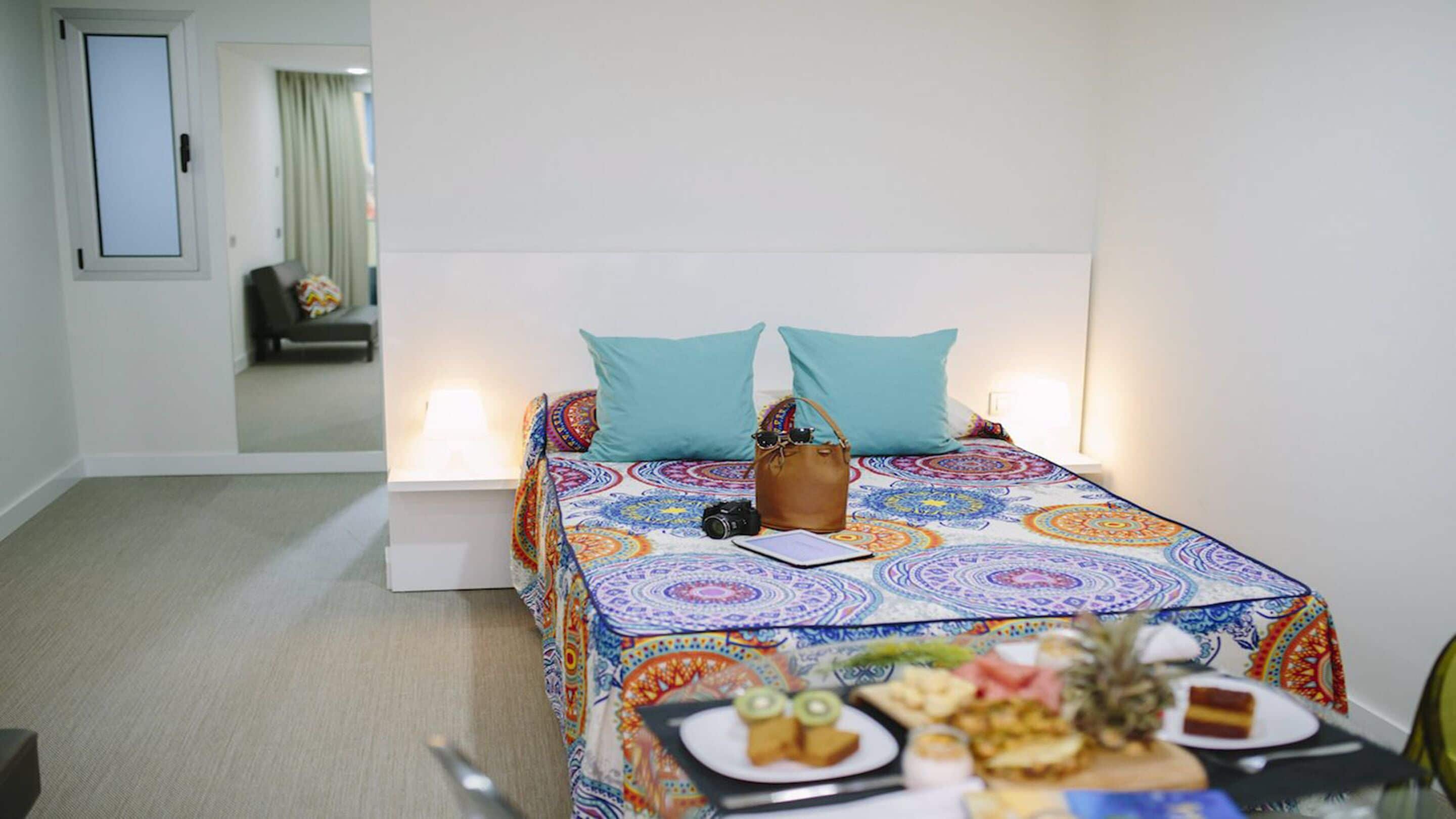 Une chambre d’hôtel avec un lit, une table de chevet avec une lampe, une petite table avec de la nourriture et un miroir qui reflète la pièce.