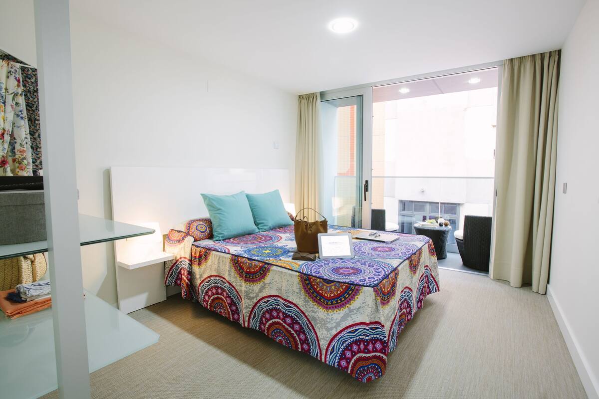 A bedroom with a bed, a bedside table, a chair, a small table, and a view of the outside.