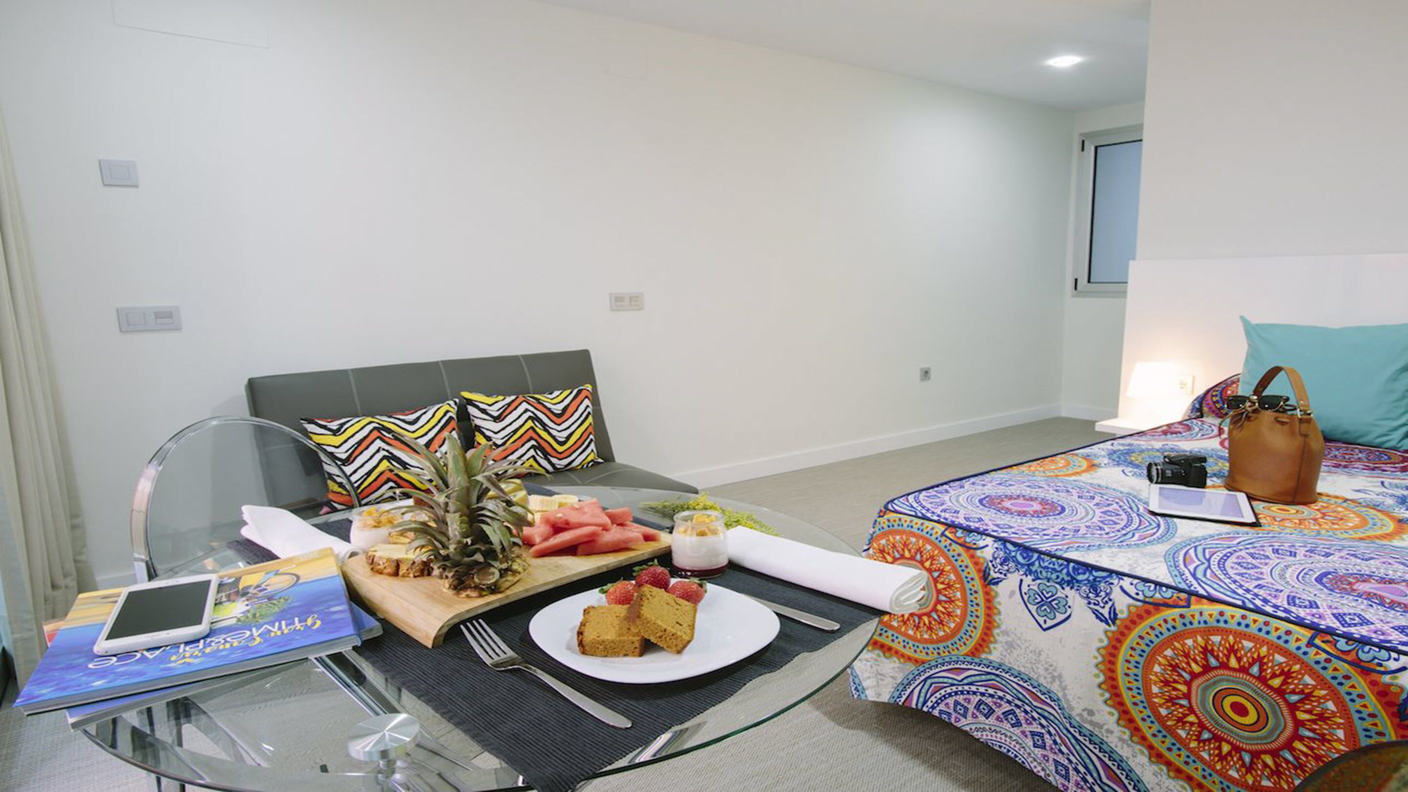 A hotel room with a bed, a desk with a laptop, a glass table with a laptop, a chair, and a small table with food and drinks.