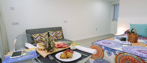 A hotel room with a bed, a desk with a laptop, a glass table with a laptop, a chair, and a small table with food and drinks.