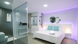 A modern hotel room with a glass shower enclosure, a white bed with teal pillows, and a wall-mounted mirror. - Valentina Beach (Las Palmas de Gran Canaria)