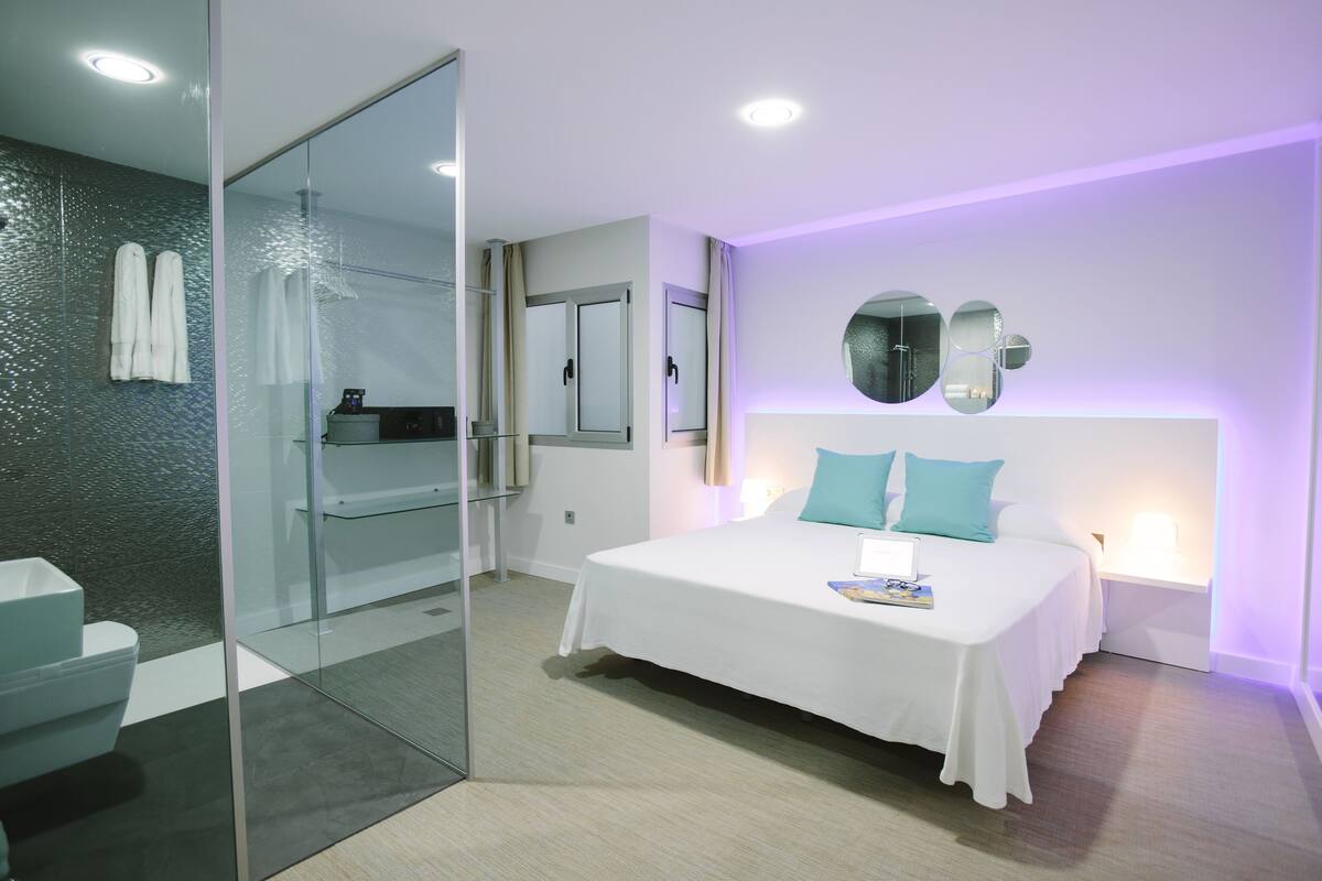 A modern hotel room with a glass shower enclosure, a white bed with teal pillows, and a wall-mounted mirror.