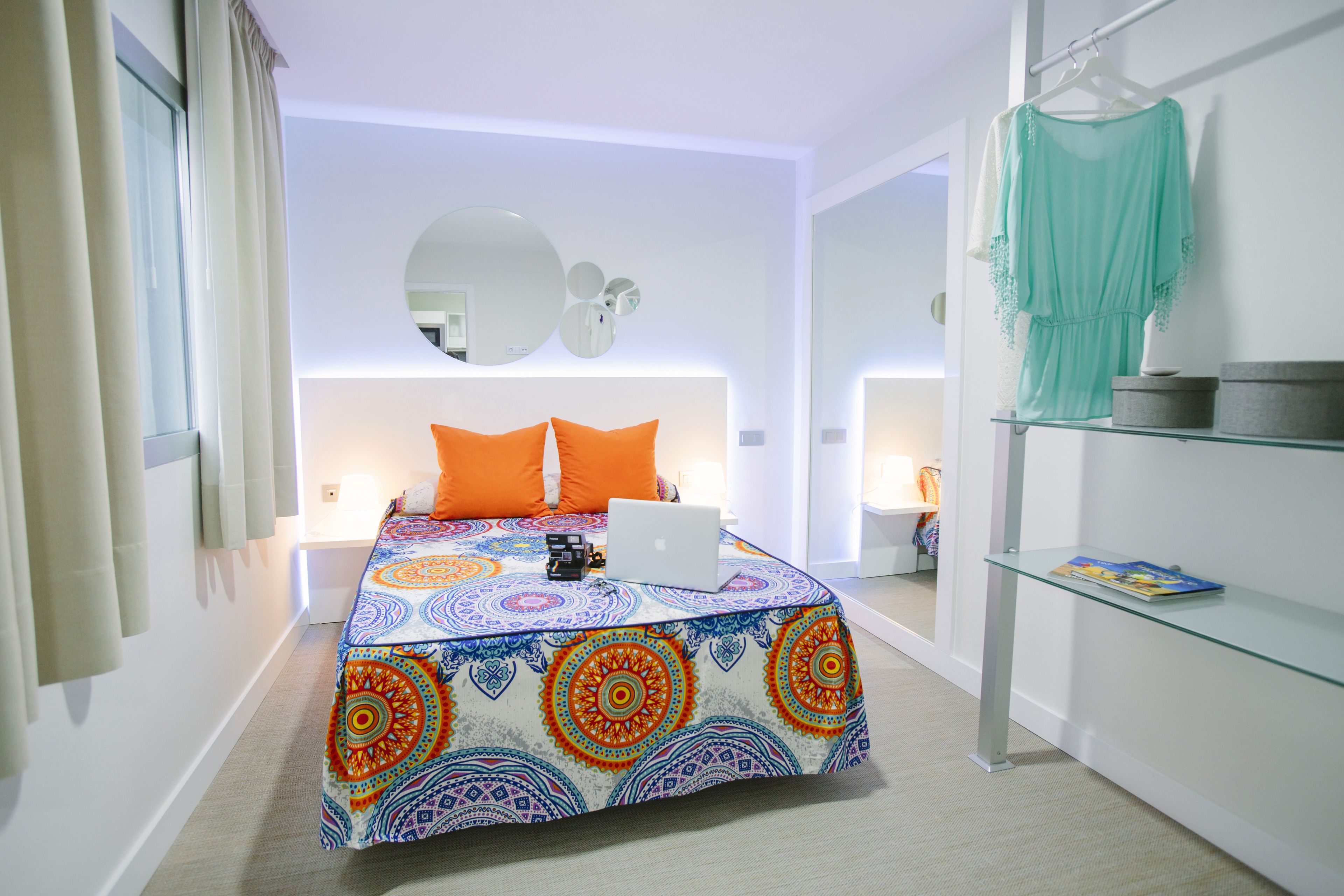 A modern bedroom with a colorful bedspread, a laptop on the bed, and a glass shelf with a book.