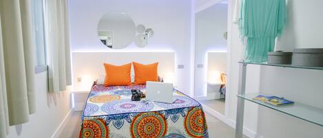 A modern bedroom with a colorful bedspread, a laptop on the bed, and a glass shelf with a book.