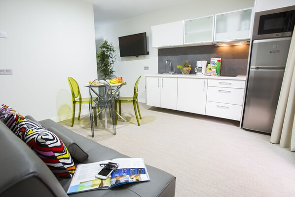 Una sala de estar moderna con un sofá gris, una mesa de comedor con sillas y una cocina con armarios blancos y un refrigerador de acero inoxidable.
