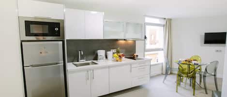 A modern kitchen with stainless steel appliances, a coffee maker, and a fruit bowl. There is a dining area with a glass table and chairs.