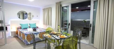 A modern hotel room with a bed, a dining table with chairs, a laptop, and a glass table.