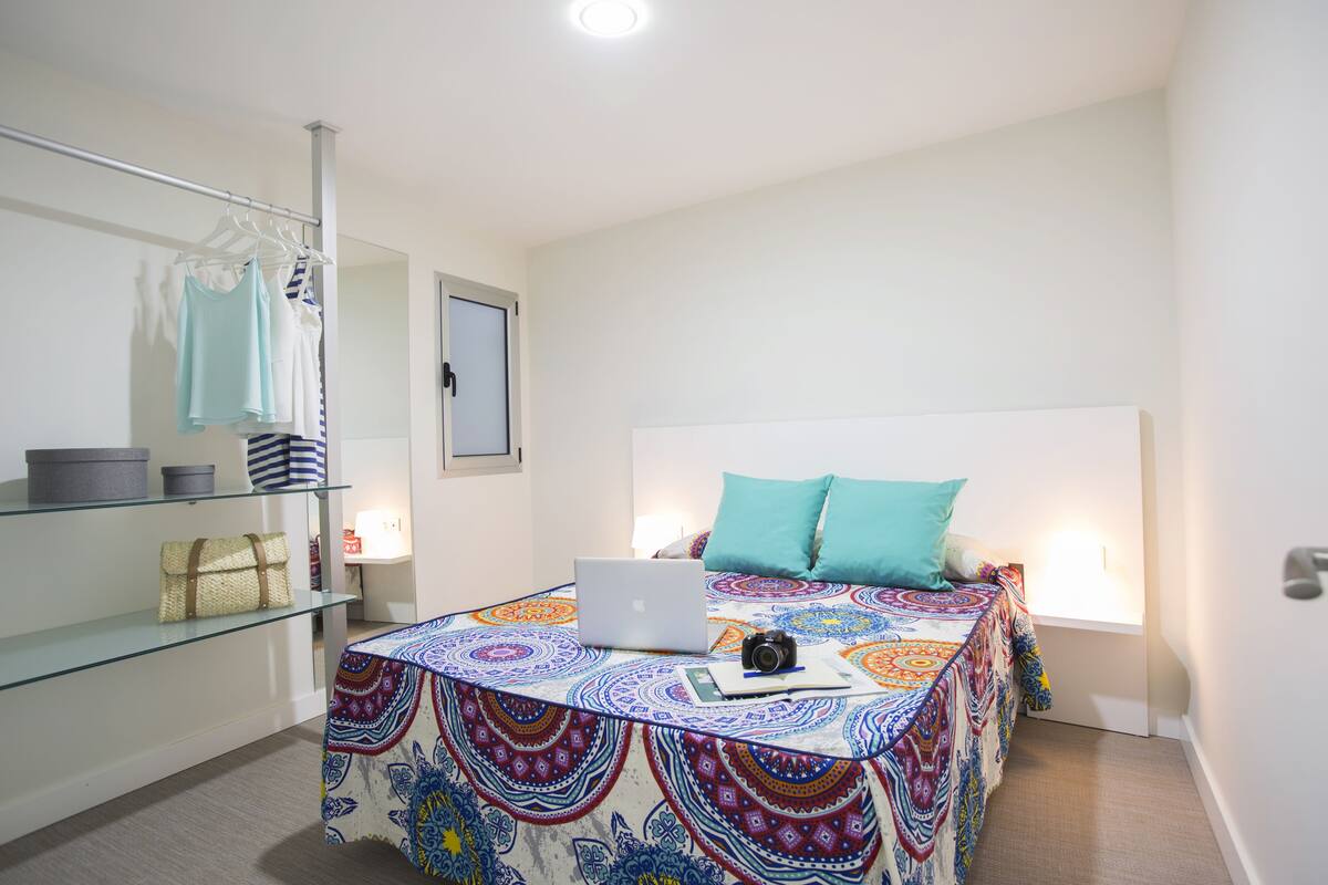 A bedroom with a bed, a laptop, a camera, and a bedside table with a lamp.