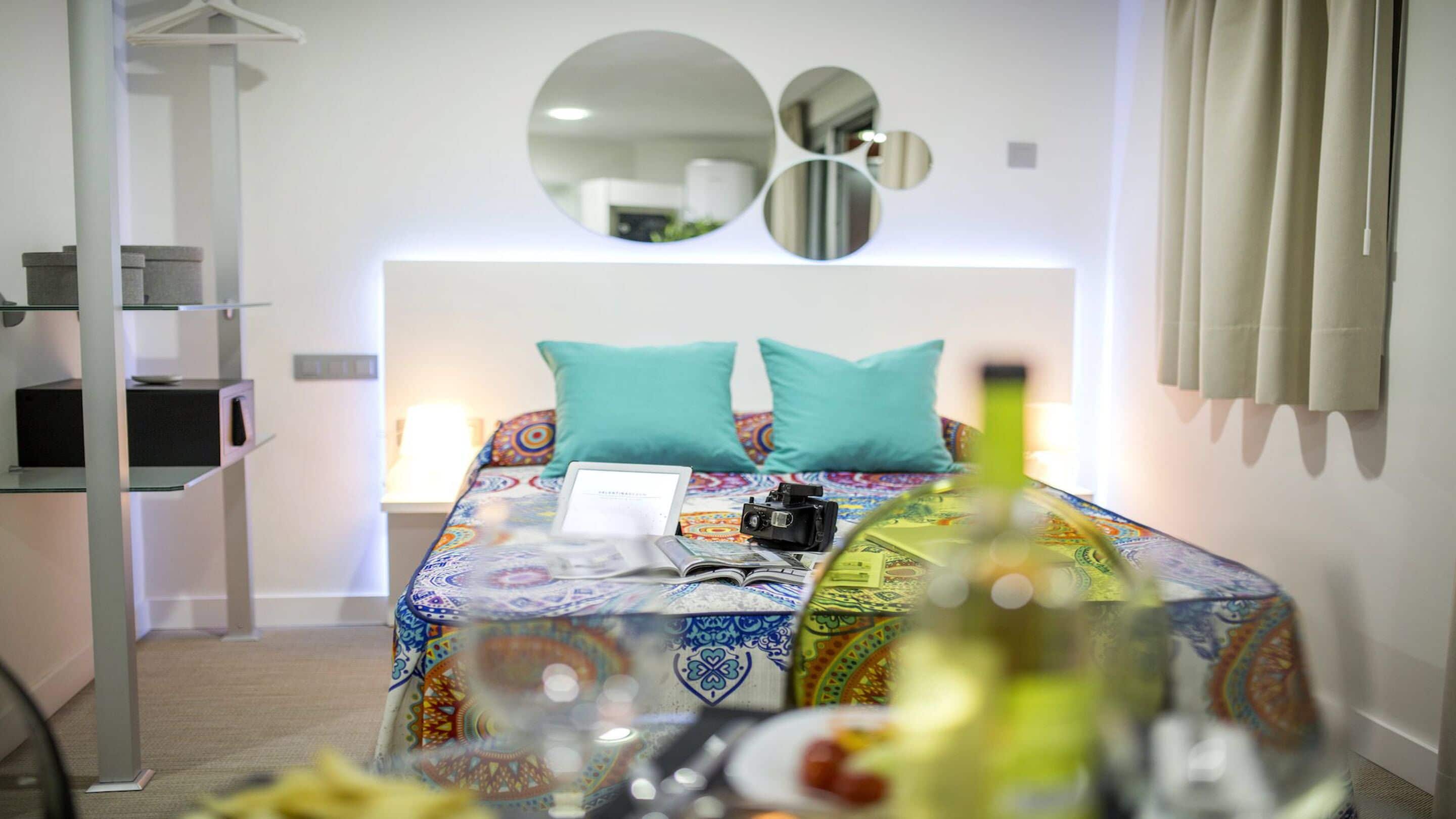 A hotel room with a bed, a laptop, a camera, and a bottle on the nightstand.