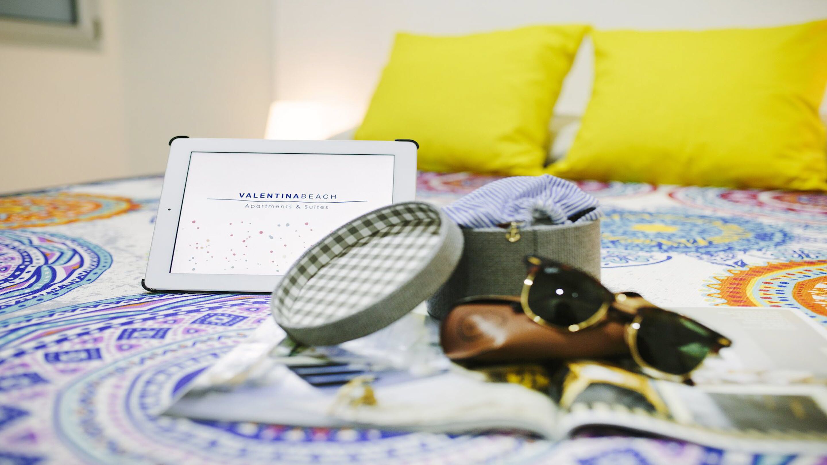 A bed with a colorful patterned bedspread, a tablet displaying 'Valentinabeach Apartments & Suites', a checkered item, a pair of sunglasses, and a small handbag.
