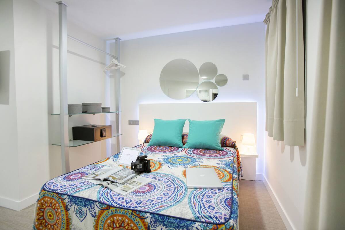 A modern bedroom with a patterned bedspread, a laptop, a camera, and a bedside lamp.