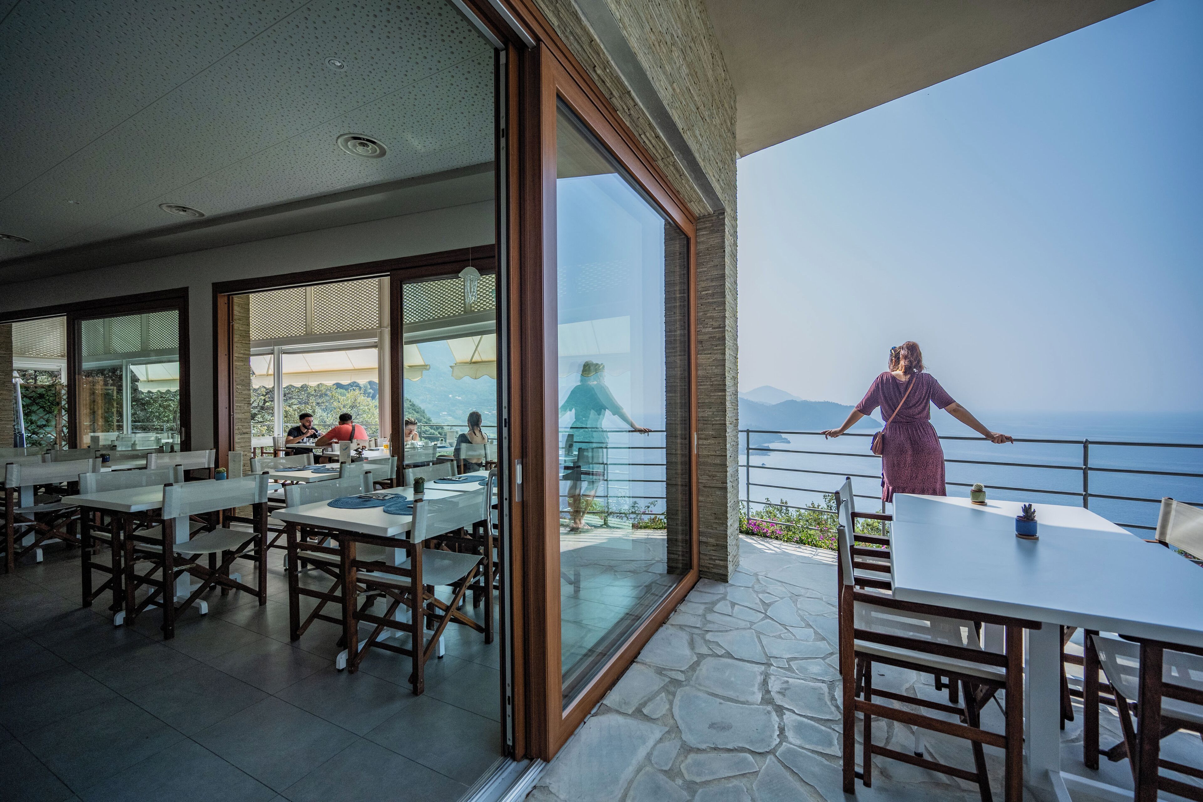 A dining area with tables and chairs, large windows, and a view of the sea.