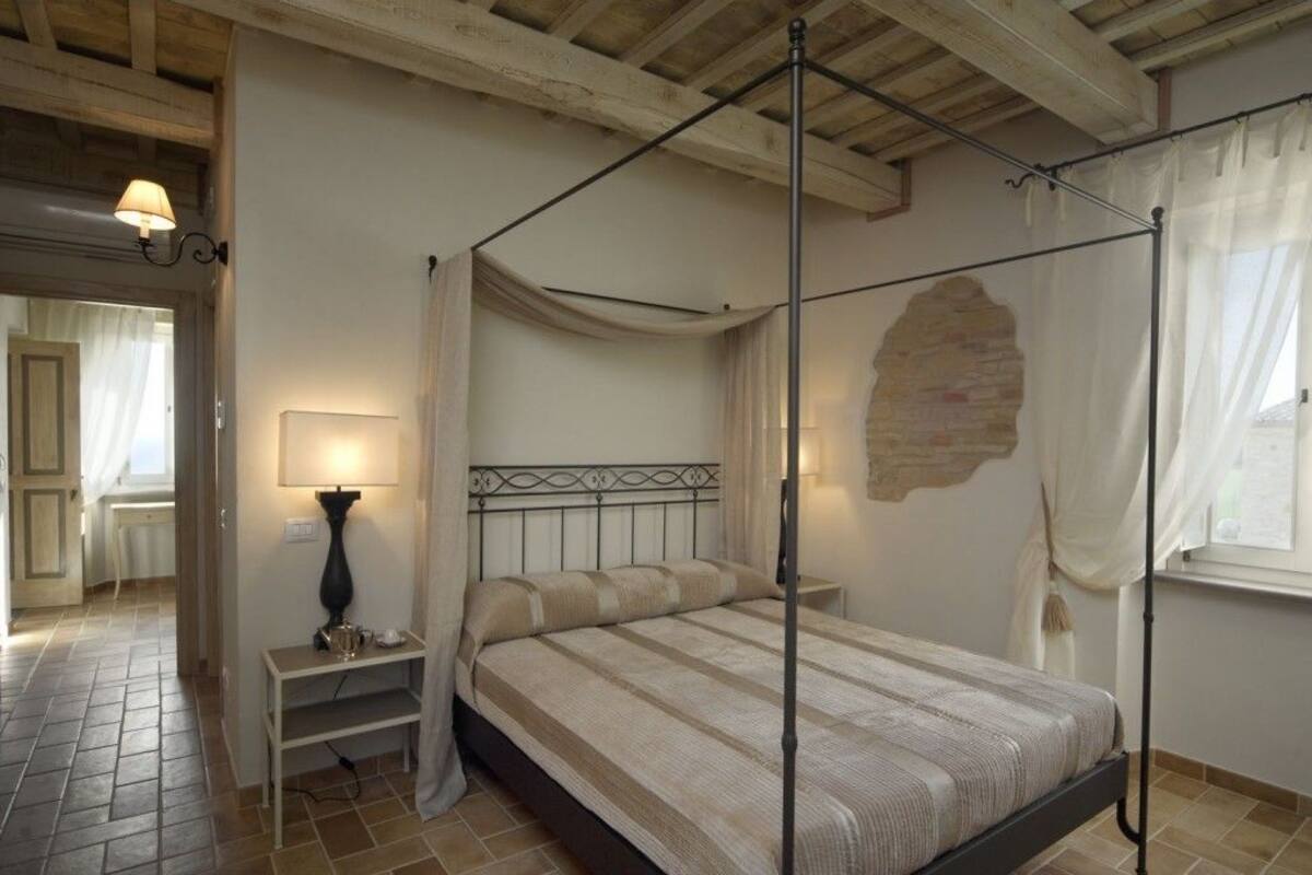 A four-poster bed in a room with a stone wall, a bedside table with a lamp, and a window with curtains.