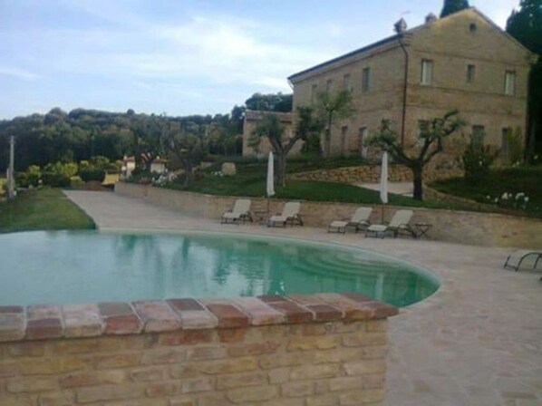 Une piscine bordée de briques, entourée d’une terrasse avec des chaises longues et un bâtiment en pierre en arrière-plan.