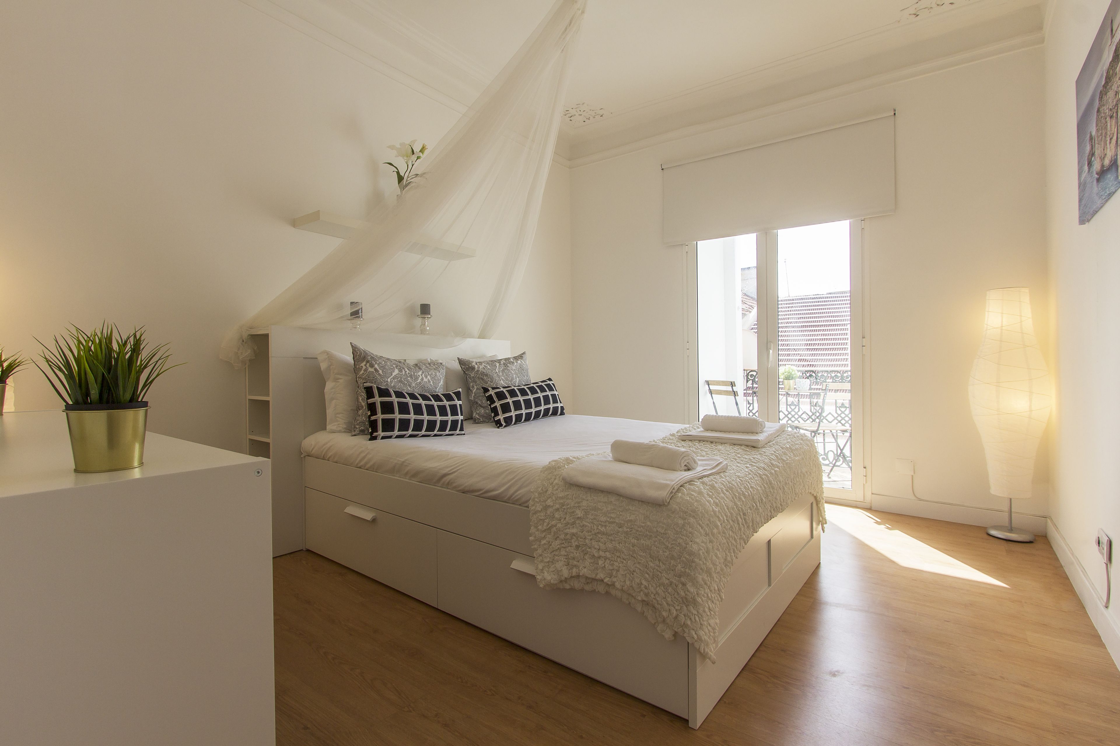 Une chambre à coucher moderne avec un lit, des tables de chevet, une plante en pot et une vue sur l’extérieur.