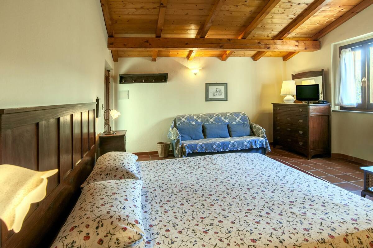 A bedroom with a wooden ceiling, a bed with floral bedding, a sofa, a dresser, and a window with curtains.