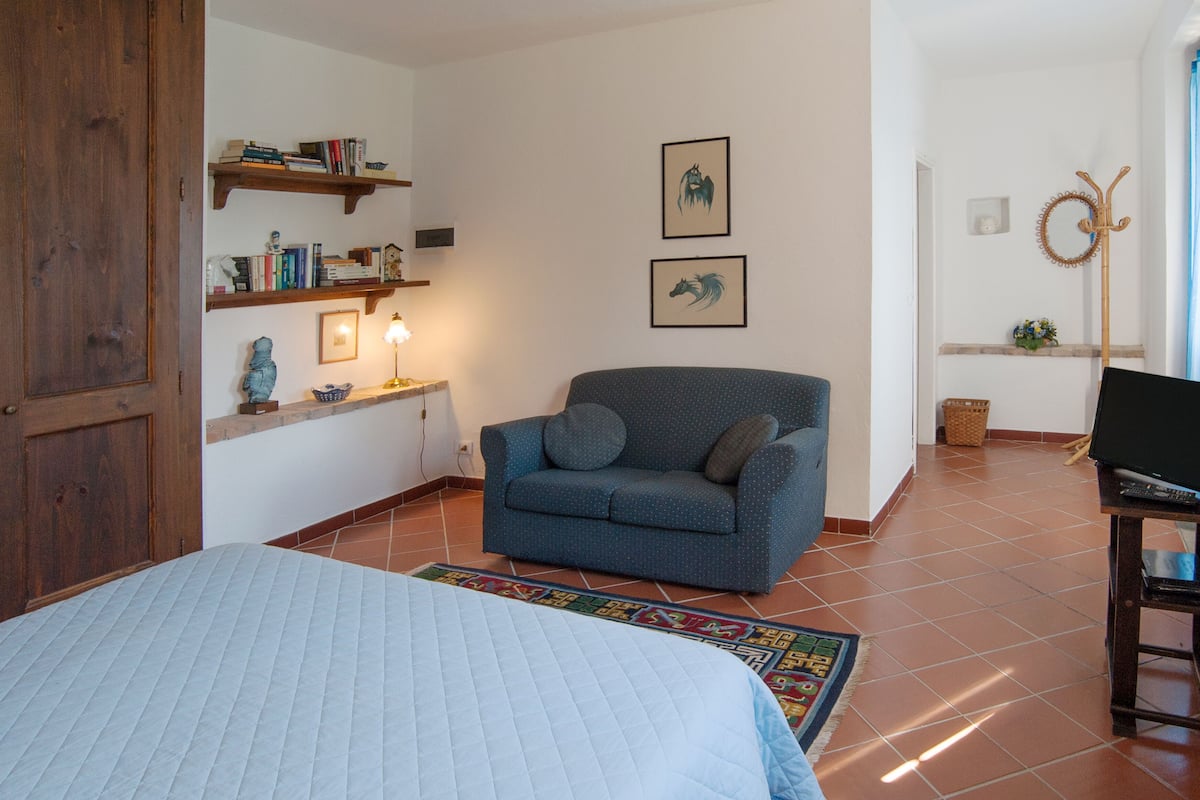 A cozy room with a bed, a blue sofa, a small table with a lamp, shelves with books, and framed pictures on the wall.