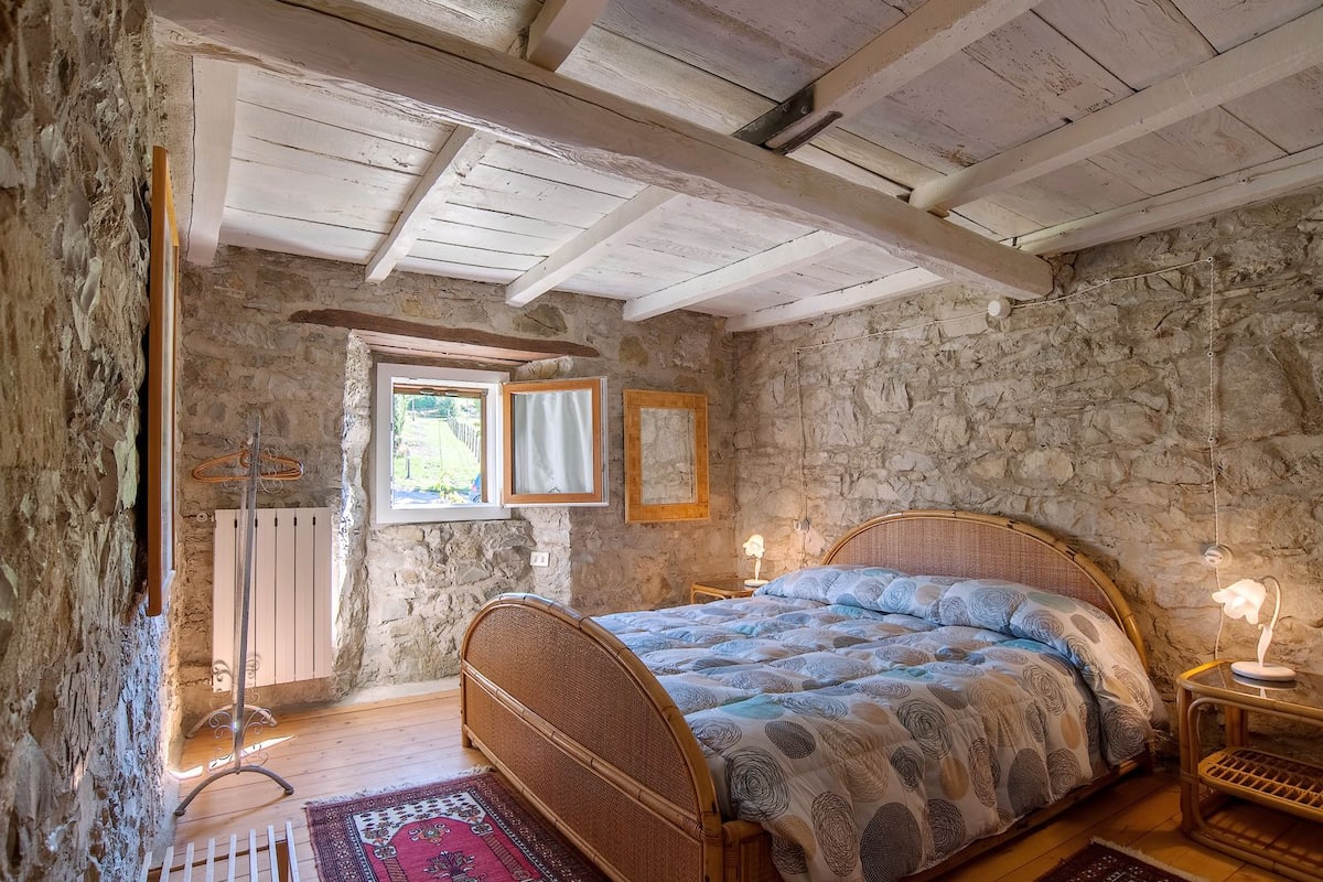 A rustic bedroom with exposed stone walls, a wooden ceiling, a single bed with a patterned bedspread, a small bedside table with a lamp, and a window with curtains.