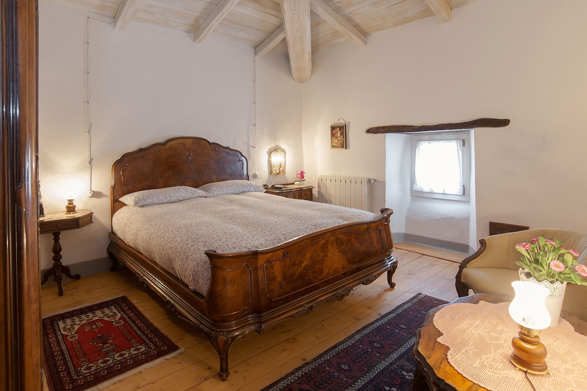 A bedroom with a wooden bed, a nightstand, a chair, a table with a lamp, a window, and a radiator.