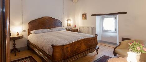 A bedroom with a wooden bed, a nightstand, a chair, a table with a lamp, a window, and a radiator.