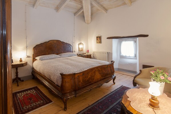 Un dormitorio con una cama de madera, una mesita de noche, una silla, una mesa con lámpara, una ventana y un radiador.