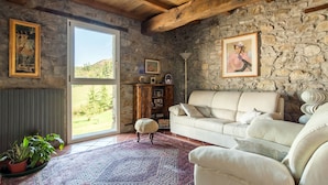 A cozy living room with a stone wall, a white sofa, a wooden ceiling, and a large window offering a view of greenery. - Fattoria Ca' di Fatino (Castiglione dei Pepoli)