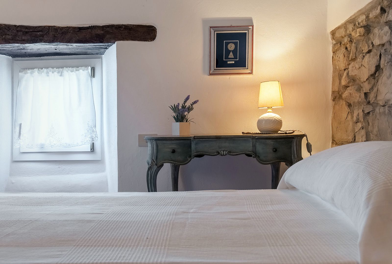 A bedroom with a bed, a nightstand with a lamp, a framed certificate on the wall, and a window with curtains.