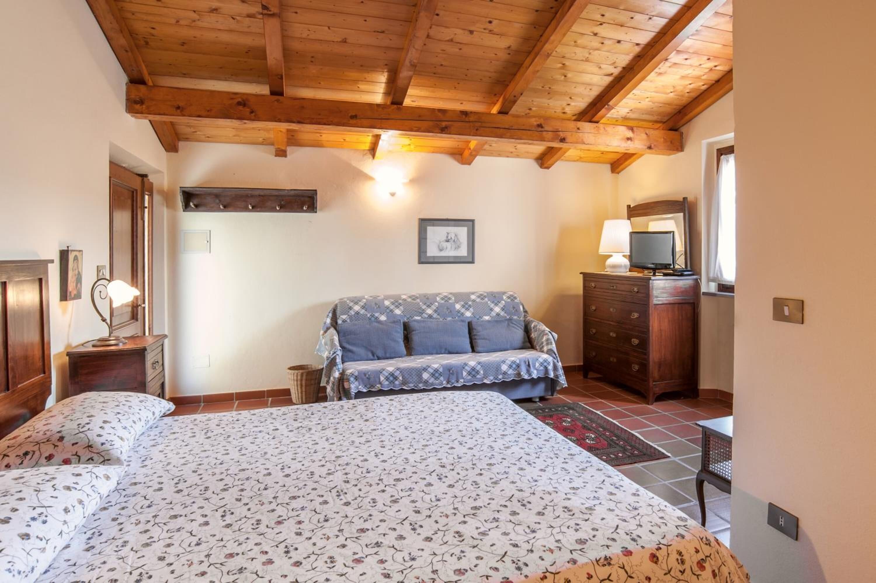 Un dormitorio con techo de madera, cama con ropa de cama floral, un sofá, cómoda y televisor.