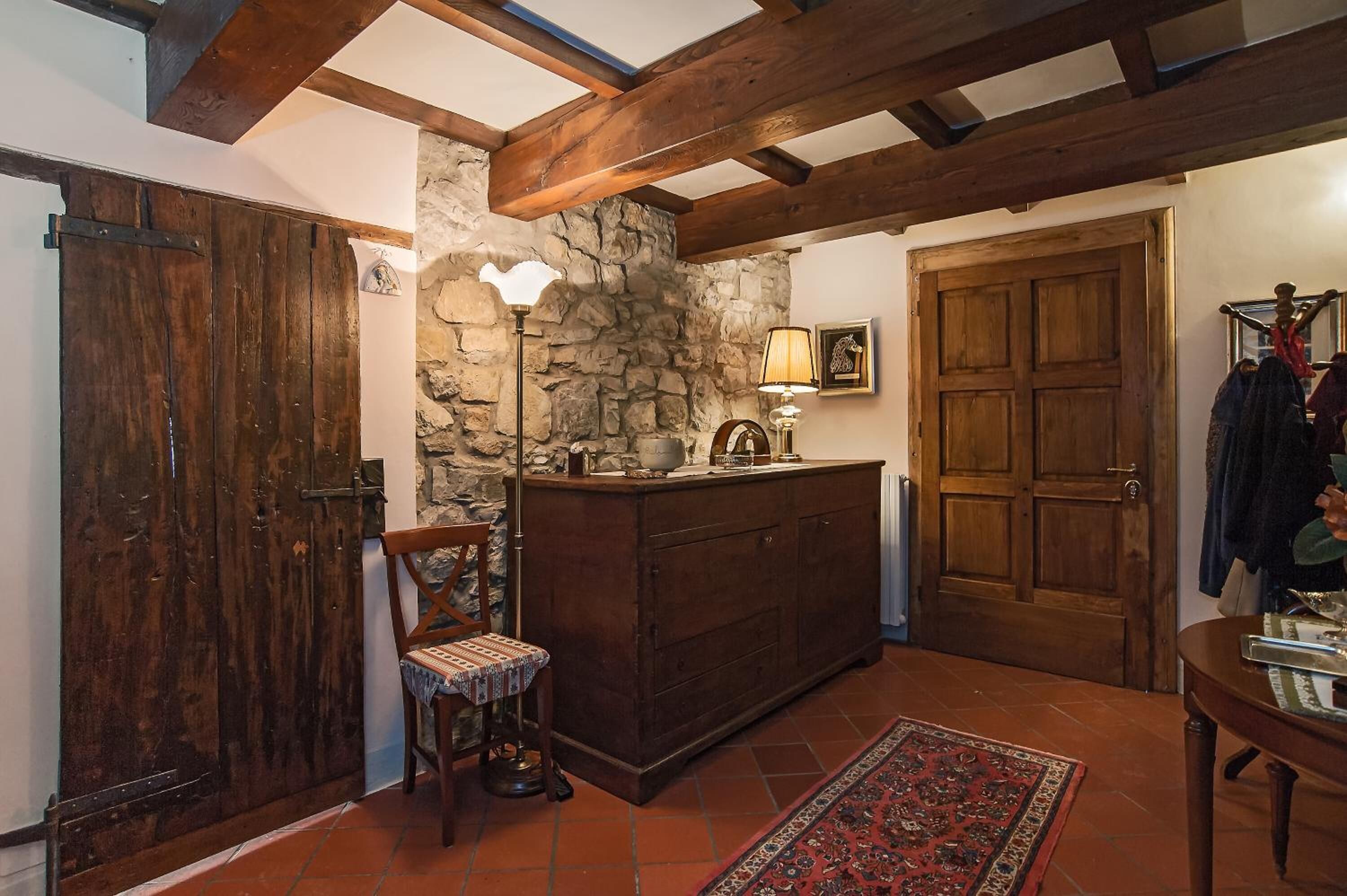 A rustic interior with exposed wooden beams, a stone wall, a wooden door, a cabinet, a chair, a table, and a floor lamp.