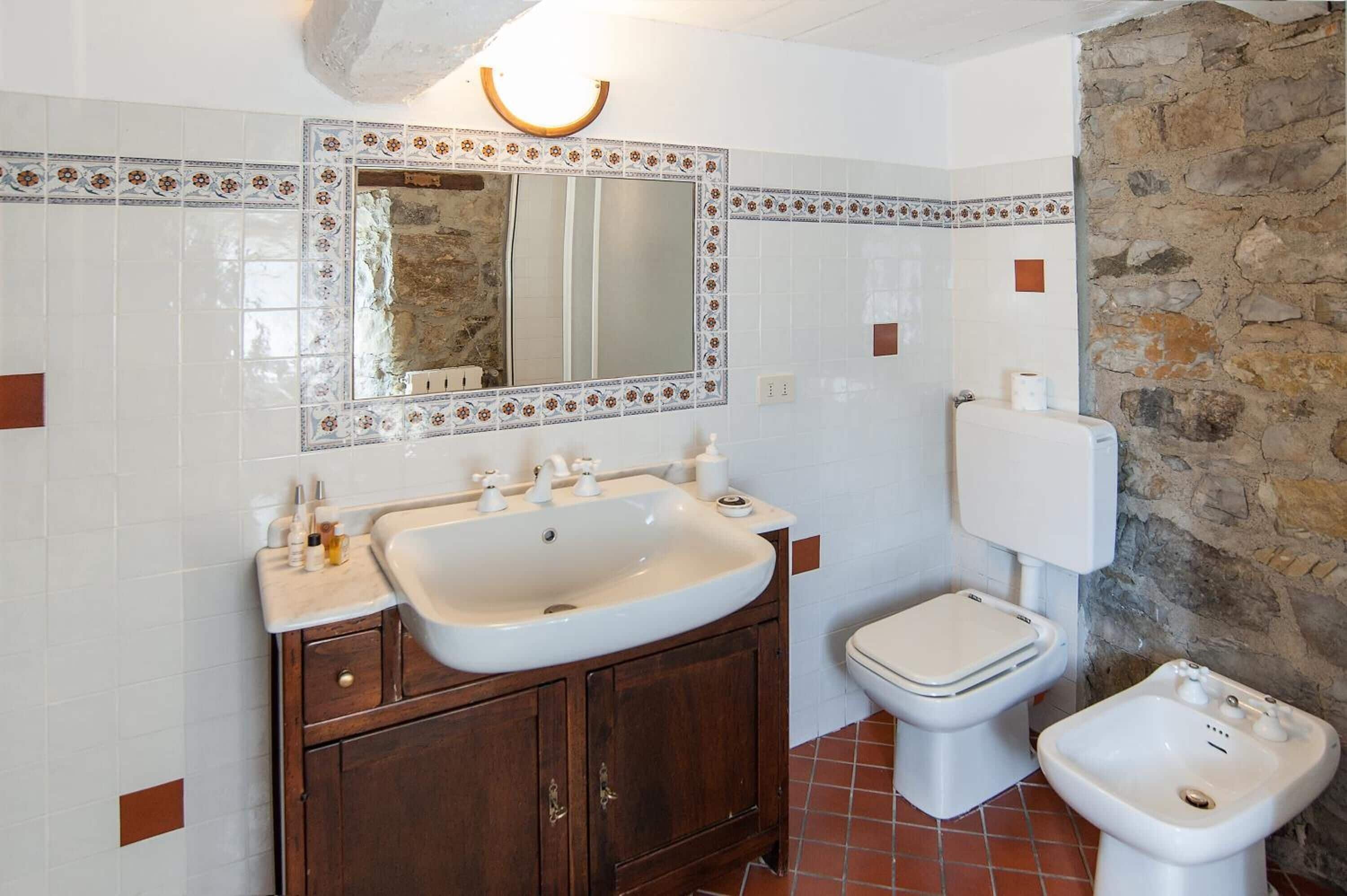 A bathroom with a white sink, wooden vanity, a mirror with decorative tiles, a toilet, and a bidet.