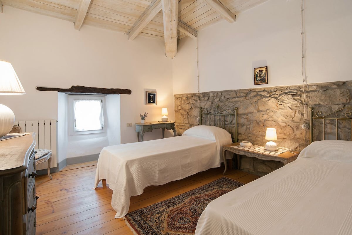 A bedroom with two single beds, a stone wall, a wooden ceiling, and a small table with a lamp.