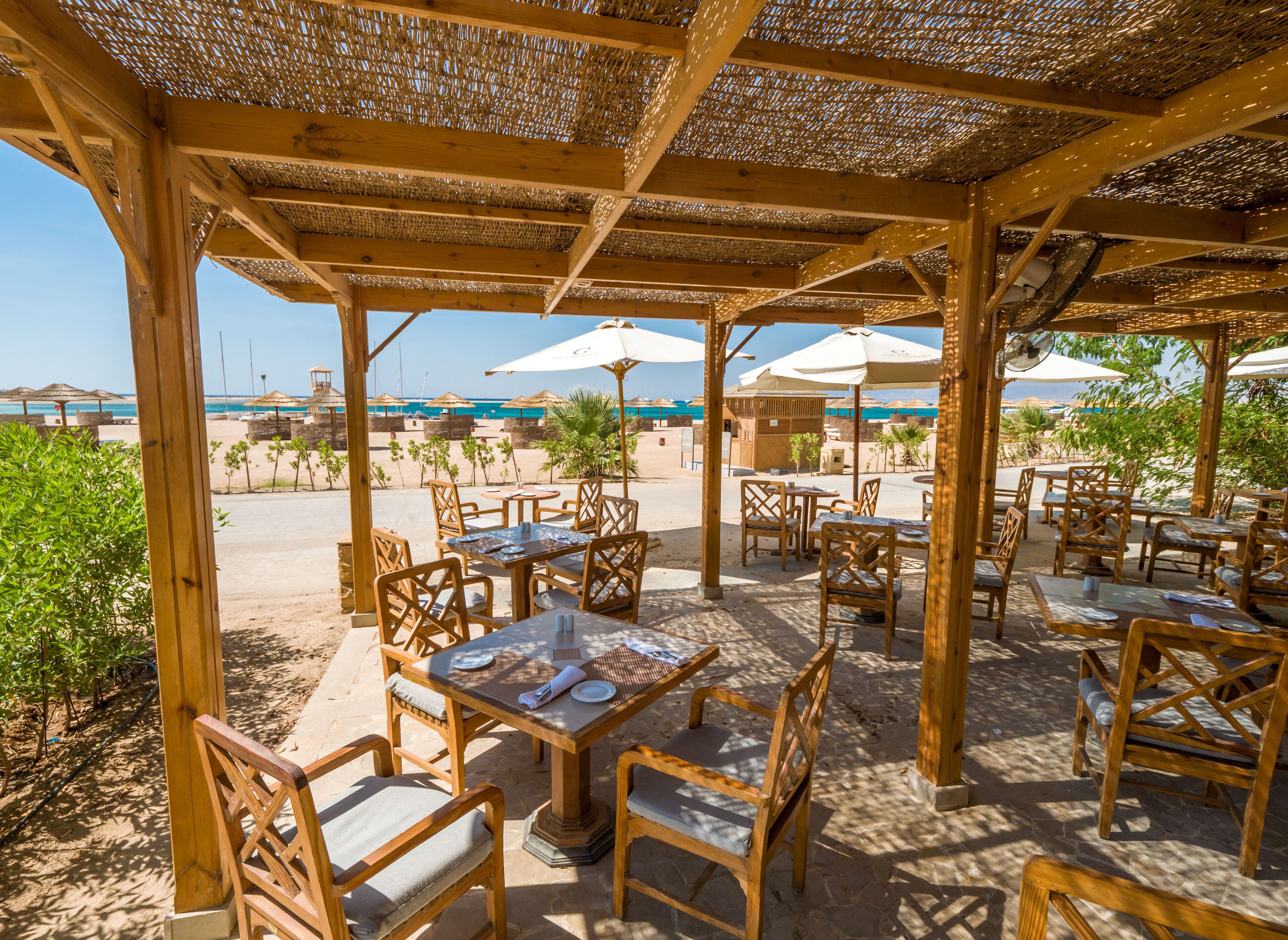Un restaurante frente a la playa con mesas y sillas de madera, sombrillas y vistas al mar.