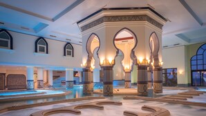 Un espace piscine intérieur luxueux, agrémenté de piliers décoratifs et d’une fontaine centrale, le tout entouré d’un vaste atrium bien éclairé.