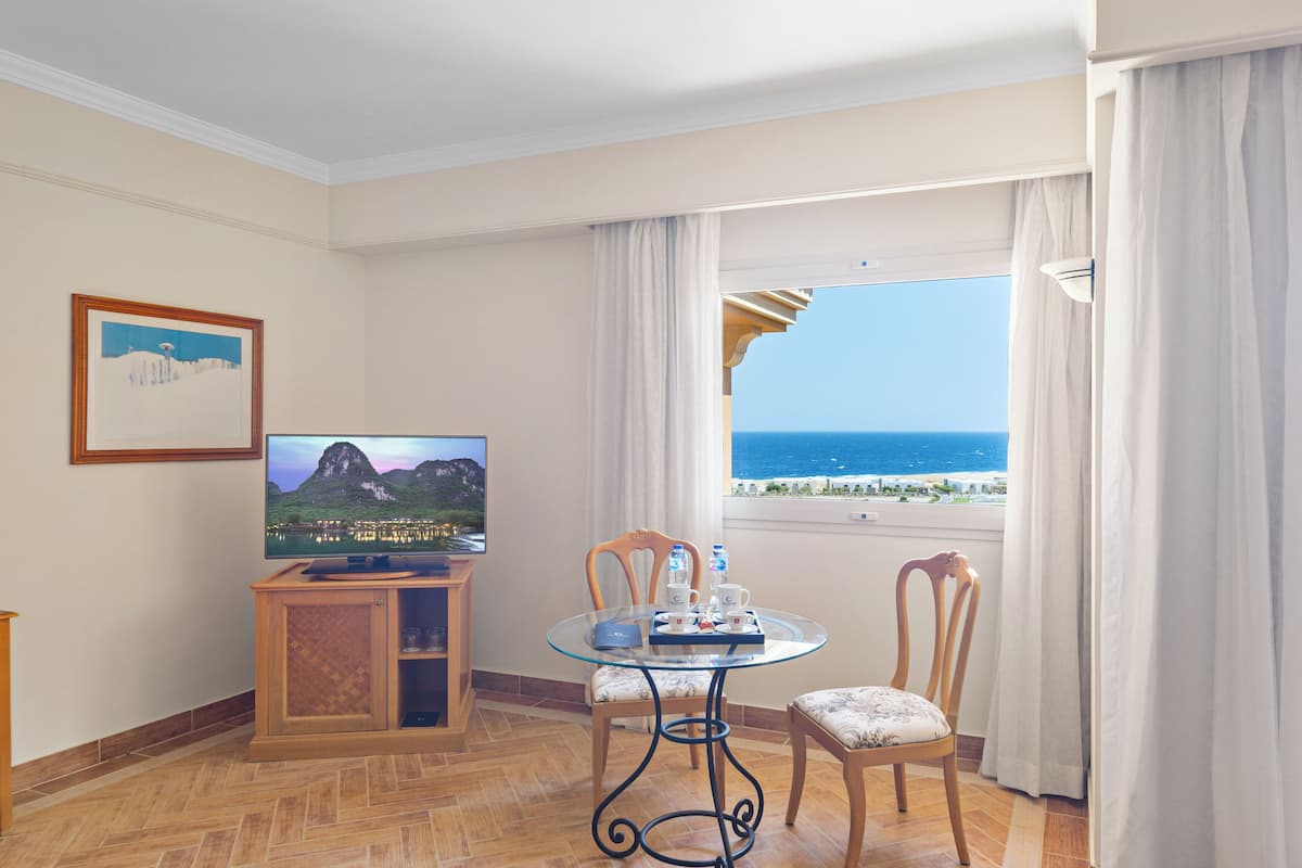 Une chambre d’hôtel équipée d’une télévision, d’une table à manger, de deux chaises, d’une fenêtre donnant sur la mer et d’un tableau encadré accroché au mur.