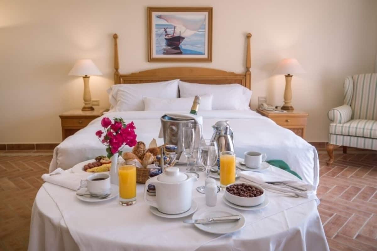 Un lit bien fait, avec du linge de lit blanc, une table de petit-déjeuner où l’on trouve des croissants, du café, du jus de fruits, et un tableau représentant un voilier accroché au mur.