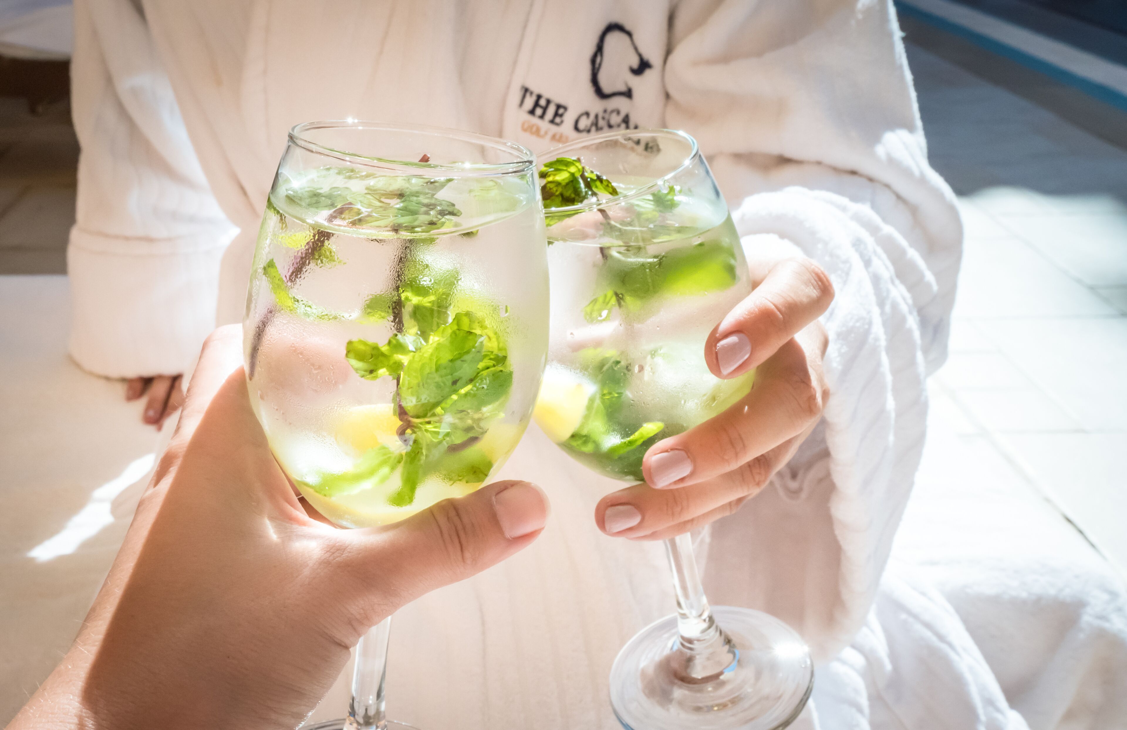 Dos personas sosteniendo vasos de una bebida transparente con hojas de menta, vestidas con túnicas blancas.