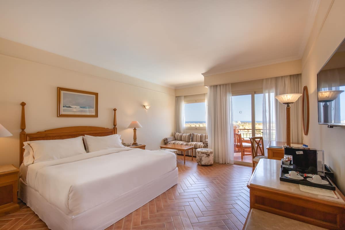 Une chambre d’hôtel avec un grand lit, un bureau avec une chaise, une petite table et une vue sur la plage par la fenêtre.