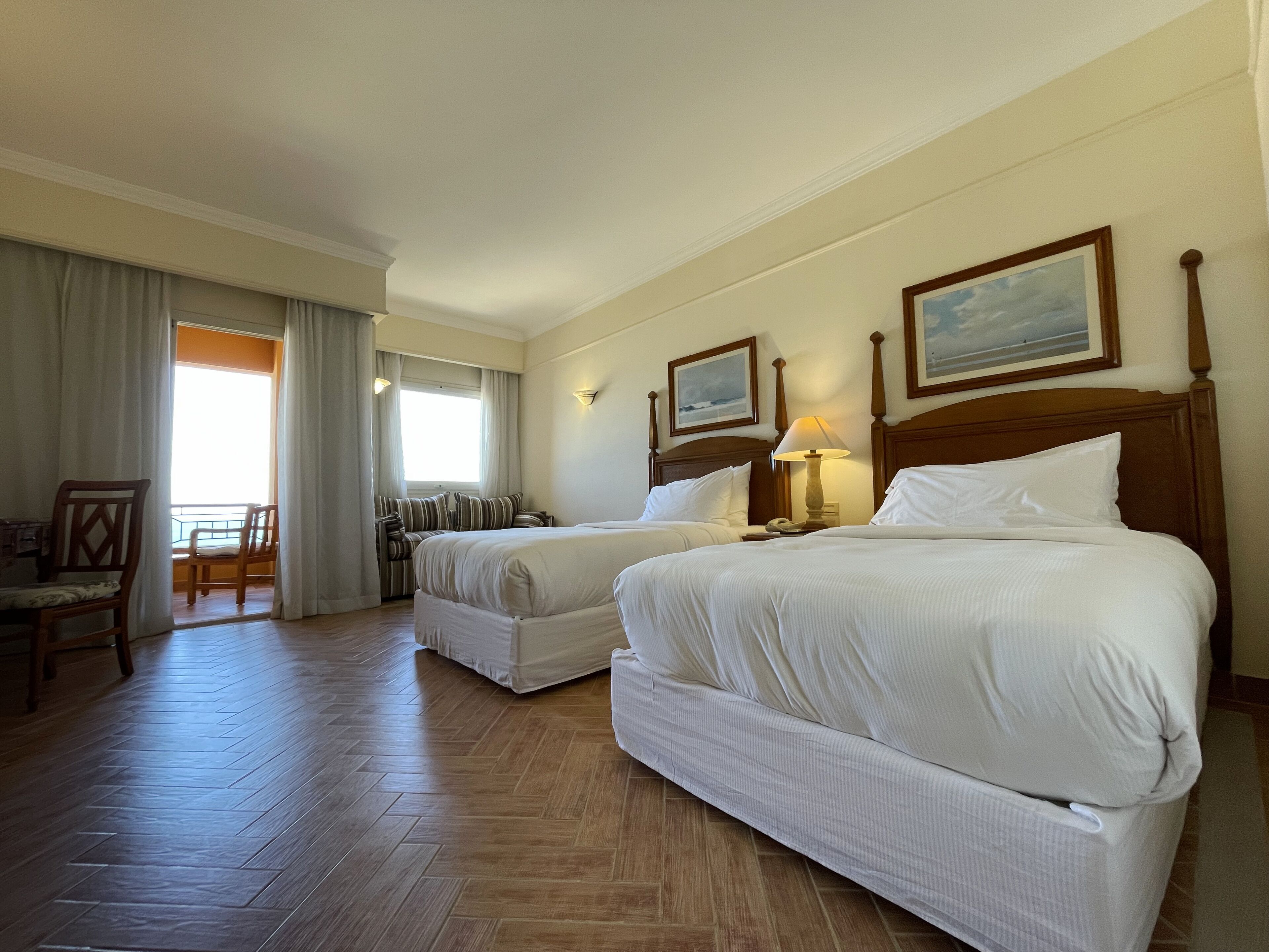 Quarto de hotel com duas camas, piso de madeira, área de refeições com mesa e cadeiras, e janela com cortinas.