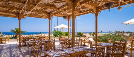 Área de refeições ao ar livre com mesas e cadeiras de madeira, estrutura de pérgola e vista para a praia e oceano.