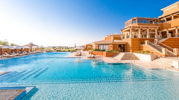 Une grande piscine entourée de chaises longues et de parasols, elle-même bordée d’un immeuble à plusieurs étages doté de balcons, le tout sous un ciel d’un bleu éclatant.