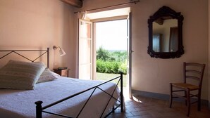 A bedroom with a bed, a chair, a mirror, and a window with an open door leading to a garden. - Agriturismo Bio Podere Doganelle (Gavorrano)