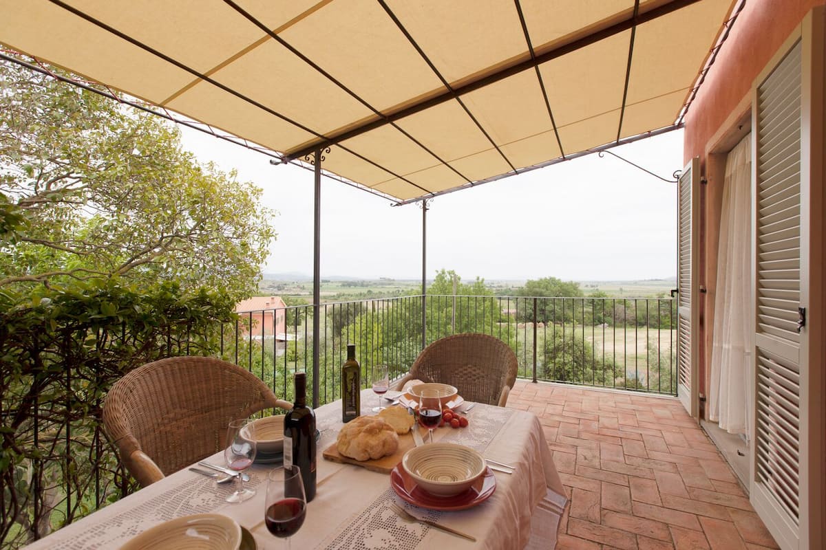 A covered outdoor dining area with a table set for two, a bottle of wine, bread, and a bowl.