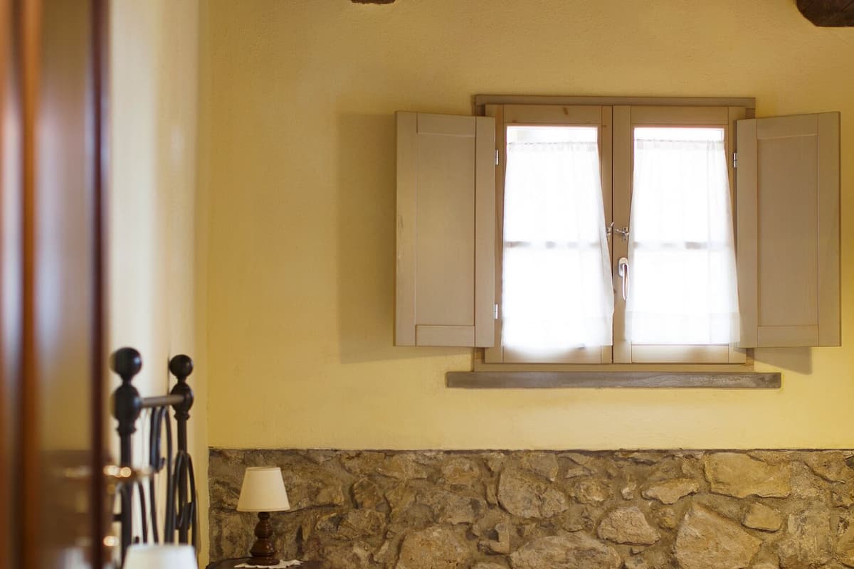 A bedroom with a stone wall, a bed with white bedding, a nightstand with a lamp, and a window with curtains.