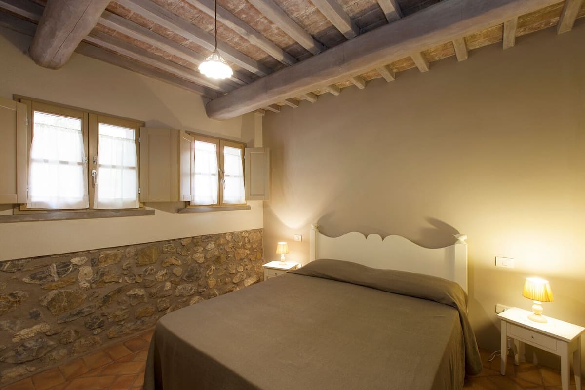 A bedroom with a stone wall, a bed with a headboard, two bedside tables with lamps, and a window with curtains.