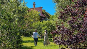 A couple walking on a grassy path surrounded by lush greenery and a building with a red roof and chimney in the background. - Agriturismo Bio Podere Doganelle (Gavorrano)