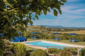 Zona de piscina con sillones de descanso y sombrillas, rodeada de vegetación y colinas al fondo.