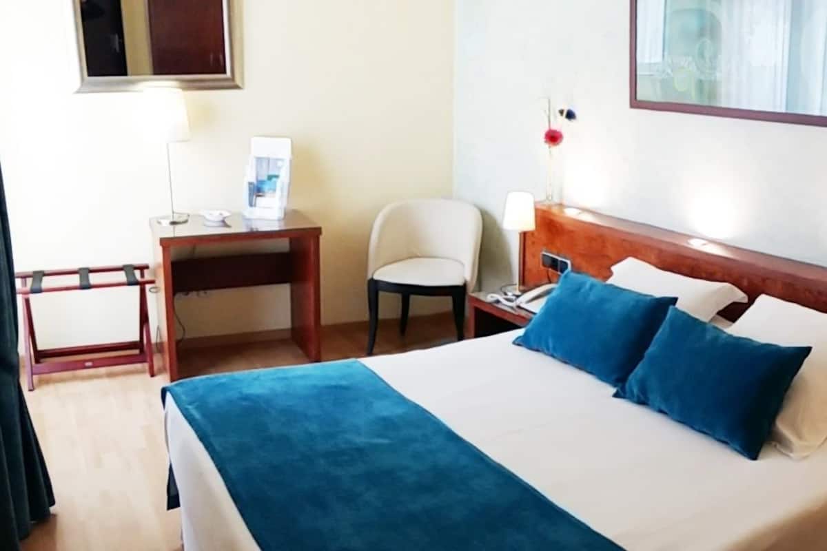 Habitació d'hotel amb llit, escriptori, cadira i tauleta de nit amb làmpada.