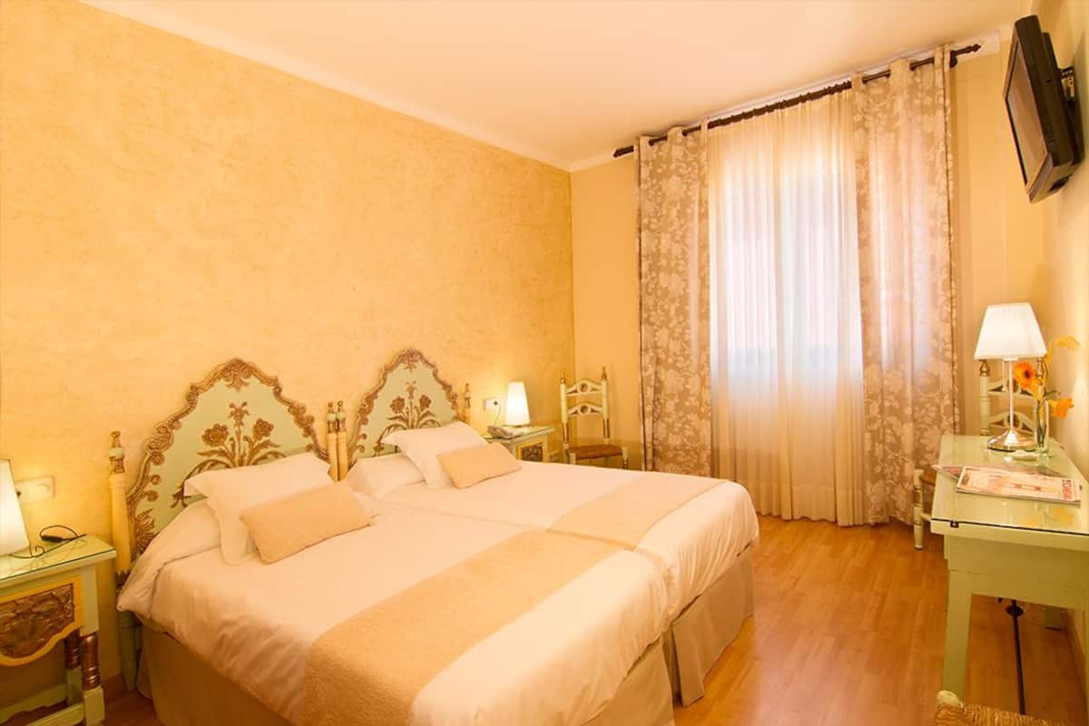 Un llit doble amb un capçal floral, dues taules de nit amb làmpades, una taula de vidre amb una làmpada i flors, i una televisió penjada a la paret.