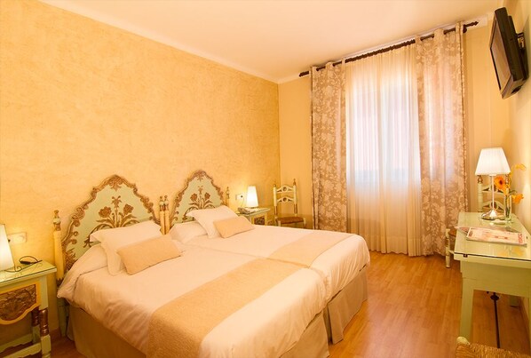 A double bed with a floral headboard, two bedside tables with lamps, a glass table with a lamp and flowers, and a television mounted on the wall. - Hotel Rosa (Begur)