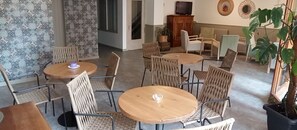 A dining area with round tables and wicker chairs, a television on a cabinet, and a potted plant. - Hotel Rosa (Begur)